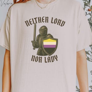 以下が含まれることがあります： ベージュ色のTシャツ。茶色で「NEITHER LORD NOR LADY」の文字がプリントされています。非バイナリープライドフラッグの色で、剣と盾を持った騎士のグラフィックが特徴です。カジュアルウェアアイテムです。