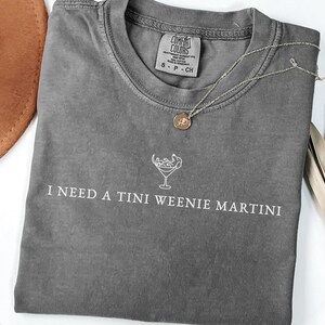 May include: Grey Comfort Colors t-shirt with the text "I NEED A TINI WEENIE MARTINI" in white. A gold necklace with a round pendant lies on the shirt. A brown hat is visible in the background.
