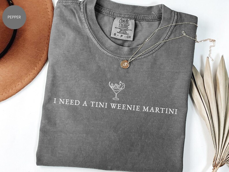 May include: Grey Comfort Colors t-shirt with the text "I NEED A TINI WEENIE MARTINI" in white. A gold necklace with a round pendant lies on the shirt. A brown hat is visible in the background.