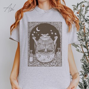 May include: A gray t-shirt with a graphic of a frog in a robe holding a cauldron with stars and a crescent moon in the background.