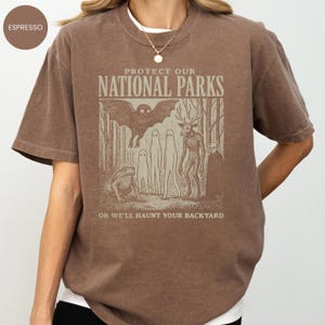 May include: Espresso-coloured t-shirt with a graphic reading "PROTECT OUR NATIONAL PARKS", featuring a bat, deer, ghosts, and a frog. The text "OR WE'LL HAUNT YOUR BACKYARD" is below. The shirt has a crew neck and short sleeves. The shirt is a brown colour.