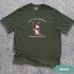 May include: An olive green short-sleeve t-shirt featuring a graphic of a corgi dressed as a wizard in a purple hat and cape, holding a wooden staff. The text above and below the dog reads OFF TO COMMIT SHENANIGANS in white capital letters.