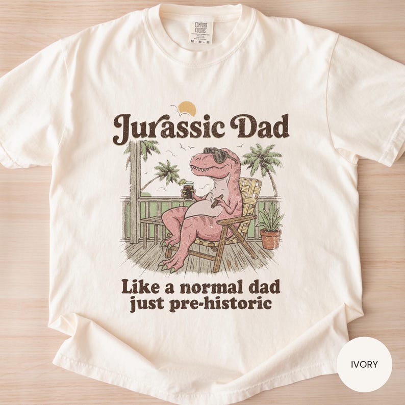 May include: Ivory-colored t-shirt with the text "Jurassic Dad" above an illustration of a relaxed dinosaur wearing sunglasses and holding a drink. The text "Like a normal dad just pre-historic" is below the illustration. The shirt is a Comfort Colors brand.
