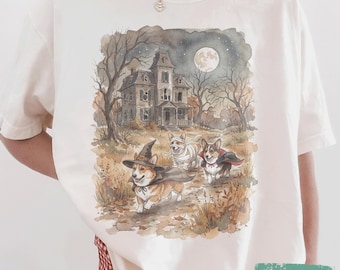 May include: An ivory t-shirt featuring a watercolour-style graphic of a haunted house under a full moon with three Corgi dogs in Halloween costumes. One dog wears a witch hat and cape, another is wrapped like a mummy, and the third wears a vampire cape.