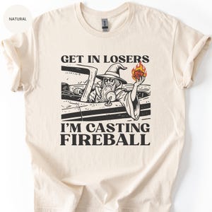 May include: Cream-colored t-shirt with a black and white graphic of a wizard driving a car, holding a fireball and a die. Text reads "GET IN LOSERS I'M CASTING FIREBALL". The shirt has short sleeves and a crew neck.