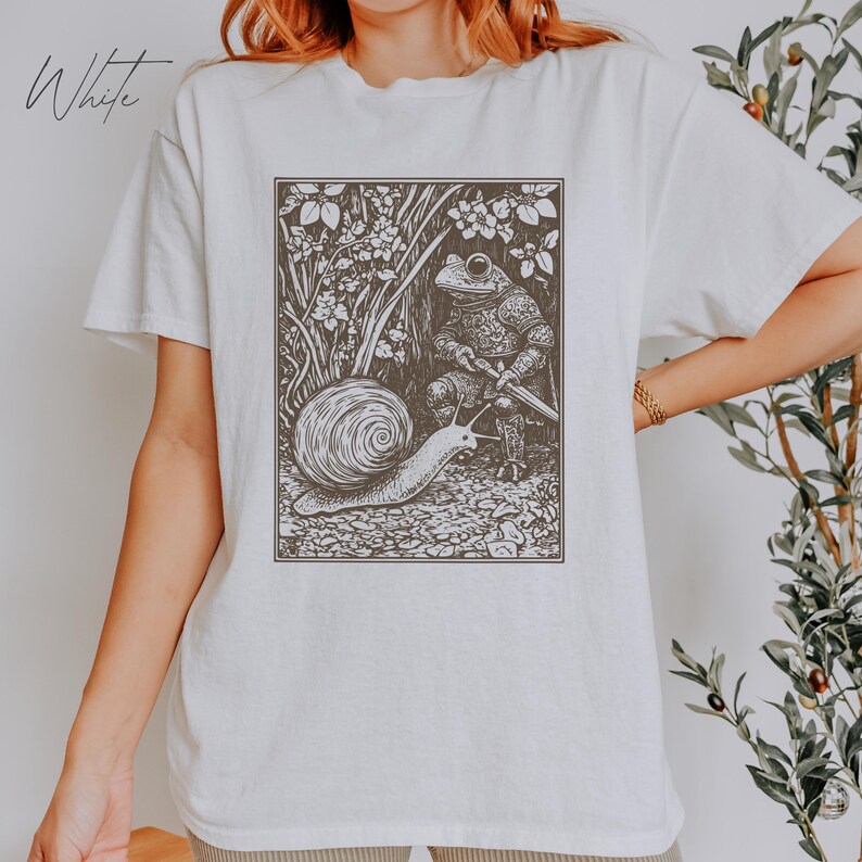 K&ouml;nnte beinhalten: Wei&szlig;es T-Shirt mit einer Schwarzwei&szlig;illustration eines Frosches in R&uuml;stung, der ein Schwert h&auml;lt, und einer Schnecke, die davonkriecht.