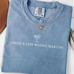 May include: A blue-jean coloured t-shirt with the phrase "I NEED A TINI WEENIE MARTINI" printed in white. A gold necklace with a pendant is draped across the shirt. The shirt is made of a soft material.