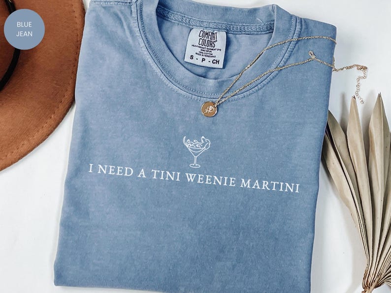 May include: A blue-jean coloured t-shirt with the phrase "I NEED A TINI WEENIE MARTINI" printed in white. A gold necklace with a pendant is draped across the shirt. The shirt is made of a soft material.