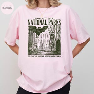 May include: Pale pink t-shirt featuring a graphic with the text "PROTECT OUR NATIONAL PARKS" above a forest scene with a bat, deer-like creature, and other figures. The text "OR WE'LL HAUNT YOUR BACKYARD" is below.