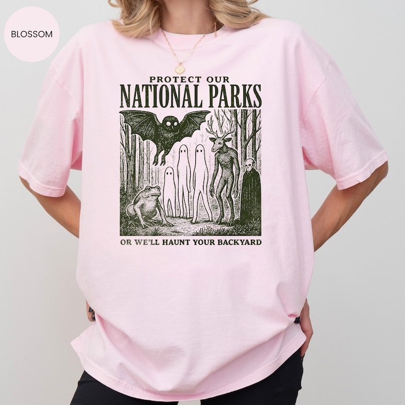 May include: Pale pink t-shirt featuring a graphic with the text "PROTECT OUR NATIONAL PARKS" above a forest scene with a bat, deer-like creature, and other figures. The text "OR WE'LL HAUNT YOUR BACKYARD" is below.