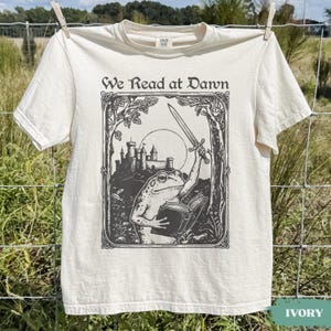 May include: An ivory short-sleeve t-shirt featuring a black graphic print of a frog holding a sword and an open book in front of a castle. The text "We Read at Dawn" is printed in a decorative font above the illustration, which is framed by trees.