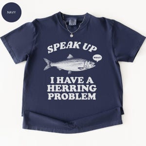 以下が含まれることがあります： 「SPEAK UP」の文字と魚のイラスト、そして「I HAVE A HERRING PROBLEM」というフレーズがプリントされたネイビーブルーのTシャツ。魚は「WHAT？」という吹き出しを持っています。Tシャツは半袖で、丸い襟ぐりです。