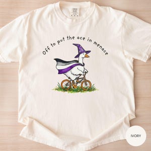 May include: Ivory-colored t-shirt featuring a cartoon goose riding a bicycle. The goose wears a wizard hat and a cape with the asexual pride flag colors. Text on the shirt reads "Off to put the ace in menace."