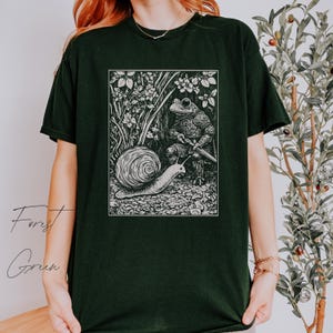 K&ouml;nnte beinhalten: Schwarzes T-Shirt mit einem wei&szlig;en Grafikdruck eines Frosches in R&uuml;stung, der ein Schwert h&auml;lt, und einer Schnecke, die auf dem Boden kriecht. Die Grafik ist in eine Waldszene mit B&auml;umen und Blumen eingebettet.