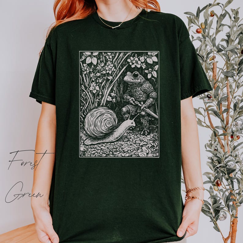 K&ouml;nnte beinhalten: Schwarzes T-Shirt mit einem wei&szlig;en Grafikdruck eines Frosches in R&uuml;stung, der ein Schwert h&auml;lt, und einer Schnecke, die auf dem Boden kriecht. Die Grafik ist in eine Waldszene mit B&auml;umen und Blumen eingebettet.