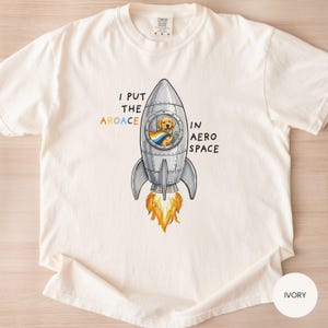 May include: Ivory-colored t-shirt featuring a cartoon rocket ship with a dog in the window. The rocket has flames and the text reads "I put the aroace in aerospace."