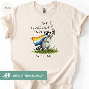 May include: Cream-colored t-shirt featuring a raccoon illustration. The raccoon, wearing a cape and holding a sword, stands on grass. Text on the shirt reads "THE BLOODLINE ENDS WITH ME."