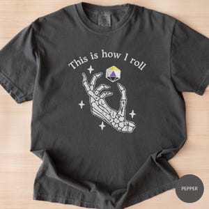 May include: Dark gray t-shirt with the text "This is how I roll" above a skeleton hand holding a multi-colored die. The design includes four white star accents, creating a unique graphic.