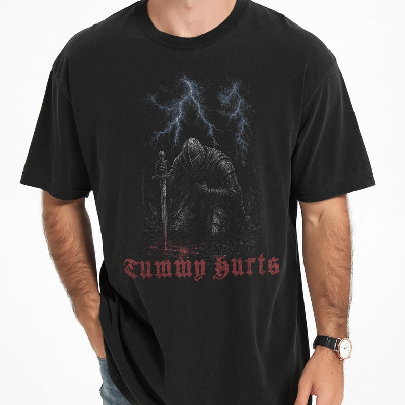 May include: A black short-sleeved t-shirt featuring a graphic of a knight kneeling with a sword under blue lightning. The text "Tummy hurts" is printed in red gothic-style lettering below the knight. The shirt has a crew neckline.