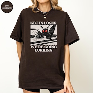 May include: Dark chocolate-colored t-shirt featuring a graphic of a moth driving a car, with the text "GET IN LOSER WE'RE GOING LURKING" in white. The design is centered on the front of the shirt. The shirt has short sleeves and a crew neck.
