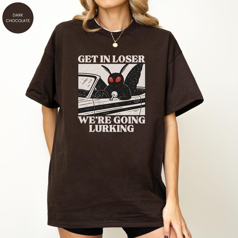 May include: Dark chocolate-colored t-shirt featuring a graphic of a moth driving a car, with the text "GET IN LOSER WE'RE GOING LURKING" in white. The design is centered on the front of the shirt. The shirt has short sleeves and a crew neck.