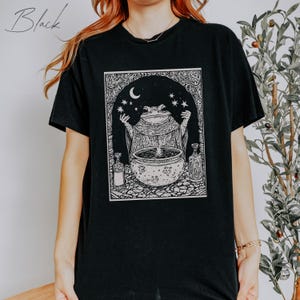 May include: Black t-shirt with a graphic print of a frog in a robe holding a cauldron with stars and a crescent moon above. The text "Black" is in the top left corner of the image.