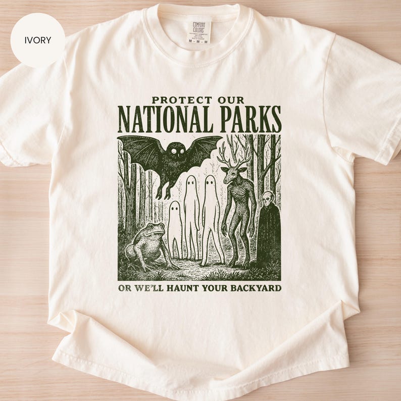May include: Ivory t-shirt featuring the text "PROTECT OUR NATIONAL PARKS" above a forest scene with cryptids. The graphic includes a bat, ghosts, a deer-like creature, a frog, and a cloaked figure. The text "OR WE'LL HAUNT YOUR BACKYARD" is below the image.