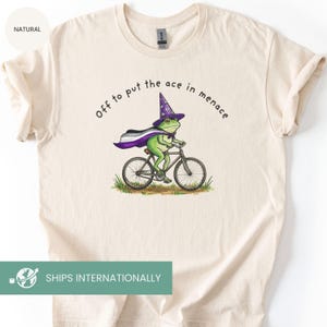 May include: Cream-colored t-shirt featuring a green frog riding a bicycle, wearing a wizard hat and cape with the asexual pride flag colors. The text "Off to put the ace in menace" is printed above the frog.