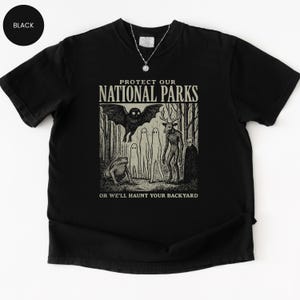 May include: Black t-shirt with a graphic design featuring forest creatures and the text "PROTECT OUR NATIONAL PARKS OR WE'LL HAUNT YOUR BACKYARD". The design includes a bat, deer-like figure, and other eerie characters.