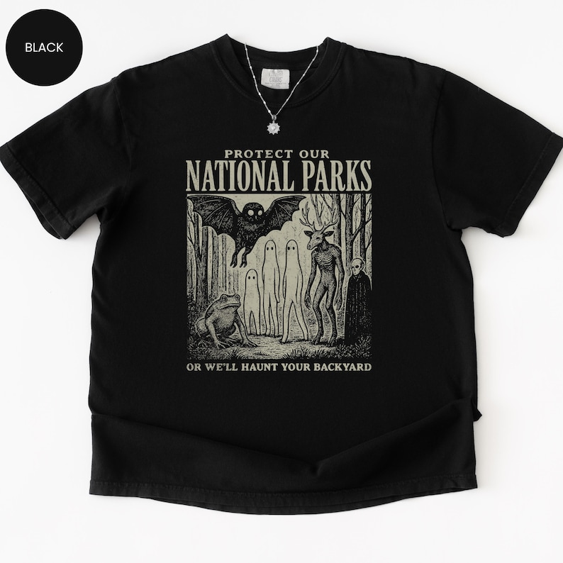 May include: Black t-shirt with a graphic design featuring forest creatures and the text "PROTECT OUR NATIONAL PARKS OR WE'LL HAUNT YOUR BACKYARD". The design includes a bat, deer-like figure, and other eerie characters.