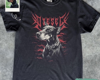 May include: A black short-sleeve t-shirt featuring a graphic of a Doberman dog wearing a collar. Above the dog is the text DIESEL in a stylised red font. The design includes a red textured background element behind the dog's head and shoulders.