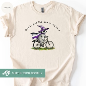 May include: Cream-colored t-shirt featuring a raccoon riding a bicycle, wearing a purple and white wizard hat and cape. The text "Off to put the ace in menace" is arched above the raccoon. The shirt has a "NATURAL" label.