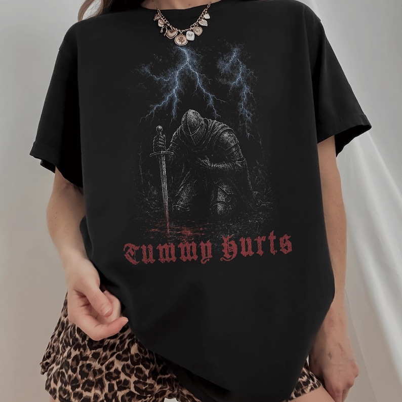May include: A black short-sleeve t-shirt featuring a graphic of a knight in armor kneeling with a sword under a lightning storm. The text "Tummy Hurts" is printed in a red gothic font below the knight. The shirt has a crew neckline.