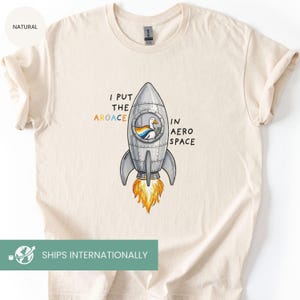 May include: Cream-colored t-shirt featuring a cartoon rocket ship with a duck inside. The rocket has the text "I PUT THE AROACE IN AERO SPACE". The rocket's flames are orange and yellow.