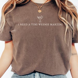 May include: Espresso-coloured t-shirt featuring the text "I NEED A TINI WEENIE MARTINI" in white, accompanied by a martini glass illustration. The shirt has a relaxed fit, ideal for casual occasions.