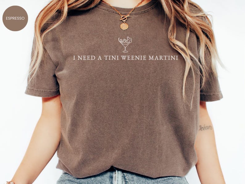 May include: Espresso-coloured t-shirt featuring the text "I NEED A TINI WEENIE MARTINI" in white, accompanied by a martini glass illustration. The shirt has a relaxed fit, ideal for casual occasions.