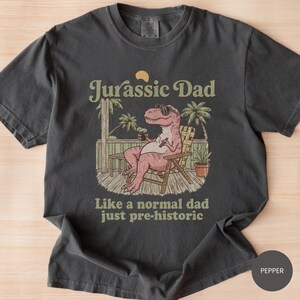 May include: Dark gray t-shirt with a graphic that reads "Jurassic Dad" above an illustration of a relaxed dinosaur wearing sunglasses and holding a drink. The text "Like a normal dad just pre-historic" is below the illustration.