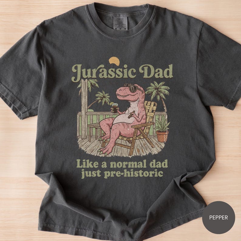 May include: Dark gray t-shirt with a graphic that reads "Jurassic Dad" above an illustration of a relaxed dinosaur wearing sunglasses and holding a drink. The text "Like a normal dad just pre-historic" is below the illustration.
