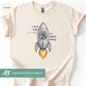 May include: Cream-colored t-shirt featuring a cartoon rocket ship with a raccoon inside, and the text "I PUT THE AROACE IN AERO SPACE". The rocket has orange flames. The shirt is made of a soft material.