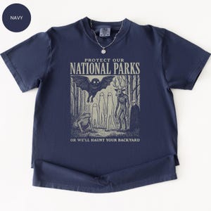 May include: Navy blue t-shirt with a graphic that reads "PROTECT OUR NATIONAL PARKS" and features a bat, frog, and other forest creatures. The text "OR WE'LL HAUNT YOUR BACKYARD" is below the graphic.