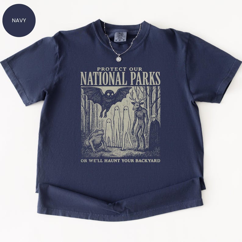 May include: Navy blue t-shirt with a graphic that reads "PROTECT OUR NATIONAL PARKS" and features a bat, frog, and other forest creatures. The text "OR WE'LL HAUNT YOUR BACKYARD" is below the graphic.