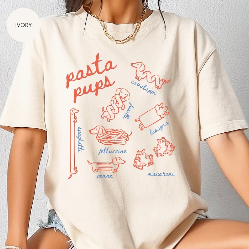May include: Ivory-colored t-shirt with a playful design featuring the text "pasta pups" in red, along with illustrations of dachshund dogs representing different pasta shapes like spaghetti, fettuccine, and lasagna. The shirt is a casual, comfortable style.