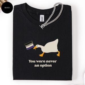 May include: Black t-shirt featuring a white goose holding an asexual pride flag. The goose has yellow feet and a yellow beak. The text "You were never an option" is printed below the goose. A silver chain is draped across the shirt.