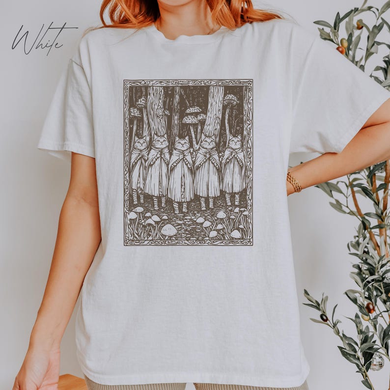May include: White t-shirt with a graphic print of five cats wearing cloaks and standing in a forest with mushrooms. The graphic is in black and white.