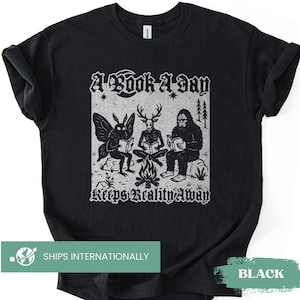 May include: A black short-sleeve t-shirt featuring a white graphic print of a moth, a deer-headed figure, and Bigfoot sitting around a campfire reading books. The text above reads A Book A Day and the text below reads Keeps Reality Away.