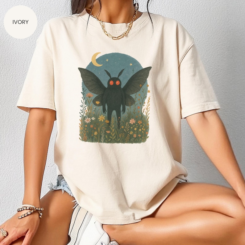 May include: Ivory-colored t-shirt featuring a whimsical illustration of a black moth with red eyes, set against a backdrop of a crescent moon and flowers. The moth has large wings and stands upright in a field of wildflowers.