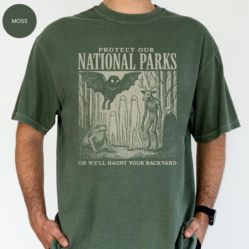 May include: Moss green t-shirt featuring the text "Protect Our National Parks" above a forest scene with cryptids. The text "Or We'll Haunt Your Backyard" is below. The shirt has a relaxed fit and is a moss green colour.