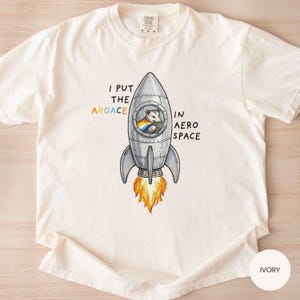 May include: Ivory-colored t-shirt with a graphic of a rocket ship and a possum in the window. The text reads "I put the aroace in aero space." The rocket has orange flames. The shirt is a Comfort Colors brand.