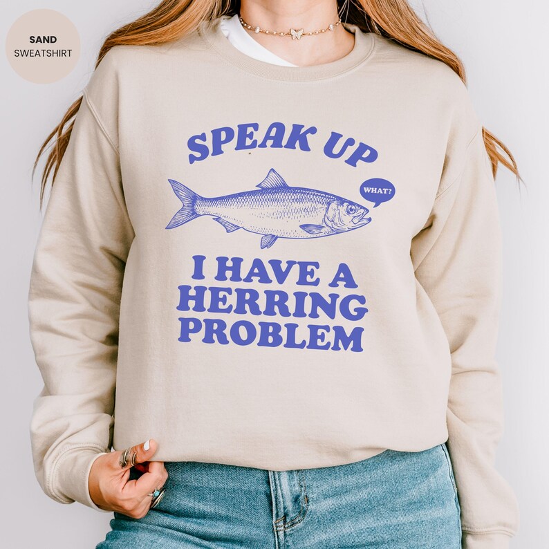 以下が含まれることがあります： 砂色のスウェットシャツ。青色の文字で「Speak Up I Have A Herring Problem」と書かれており、ニシンのイラストが描かれています。クルーネックで、様々な服装に合うニュートラルカラーです。