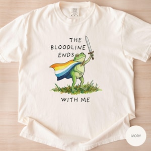 May include: Ivory-colored t-shirt featuring a green frog illustration holding a sword and wearing a cape. The text "THE BLOODLINE ENDS WITH ME" is printed above and below the frog graphic. The shirt is made of a soft material.